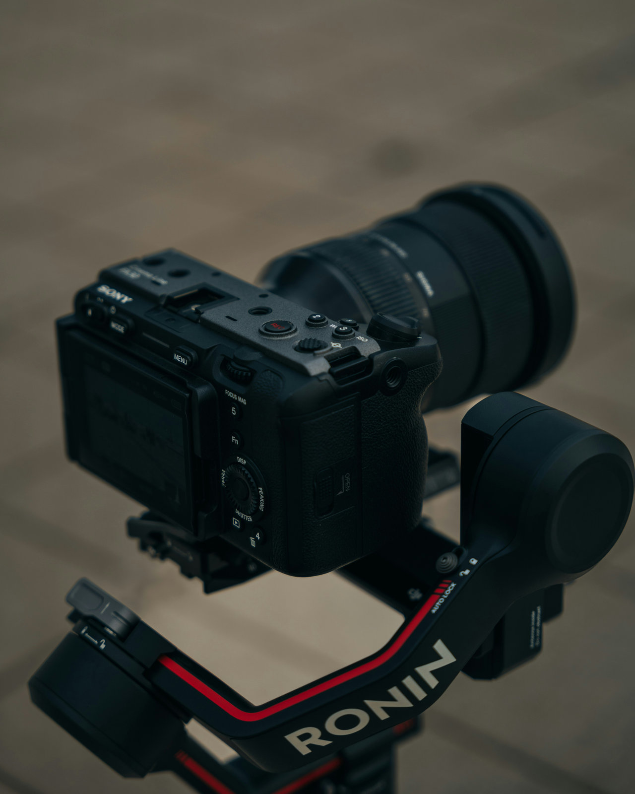 A camera mounted on a Ronin gimbal in a neutral setting. The camera has a lens attached, and the gimbal shows red and black accents.